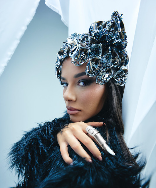 TAYNA wearing a silver sculptural headpiece and a dark feathered outfit